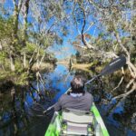 Person kayaking on a calm, narrow waterway surrounded by trees and clear blue sky, with reflections visible on the water.