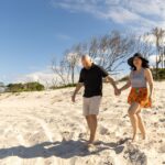 A man and woman walk barefoot on a sandy beach, holding hands and smiling, with sparse trees and blue sky in the background.