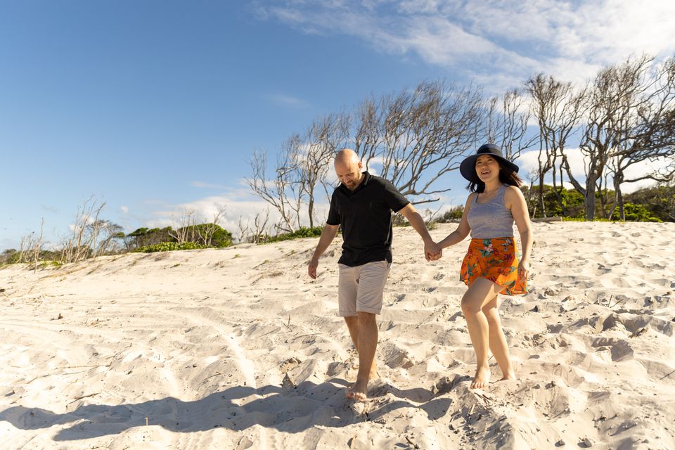 A man and woman walk barefoot on a sandy beach, holding hands and smiling, with sparse trees and blue sky in the background.