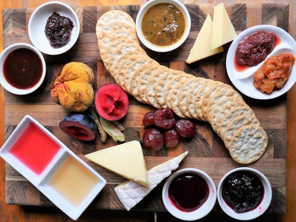 A wooden board with crackers, assorted cheeses, sliced sausages, fresh and dried fruits, and small bowls of jams, jellies, and sauces arranged neatly.