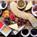 A wooden board with crackers, assorted cheeses, sliced sausages, fresh and dried fruits, and small bowls of jams, jellies, and sauces arranged neatly.