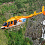 A yellow and white Zoom Helicopter flies over a green landscape with a waterfall and a train visible in the background.