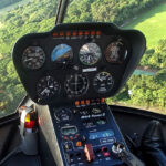 Close-up view of a helicopter cockpit with control panels and gauges, flying above a green landscape with trees and open fields visible through the windshield.