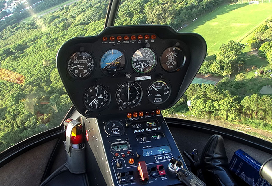 Close-up view of a helicopter cockpit with control panels and gauges, flying above a green landscape with trees and open fields visible through the windshield.