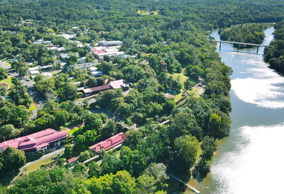 Aerial view of a small town with buildings surrounded by dense greenery, adjacent to a river with a bridge crossing over it.