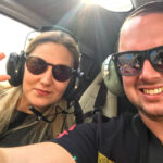 Two people wearing sunglasses and headsets sit inside a helicopter; one is making a peace sign, both are smiling.
