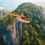 An orange and white helicopter with "Zoom" branding flies over a lush green mountainous landscape with rocky cliffs and trees.