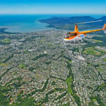 A yellow helicopter flies over a coastal city with dense green areas, residential neighborhoods, and distant mountains near the shoreline under a clear sky.