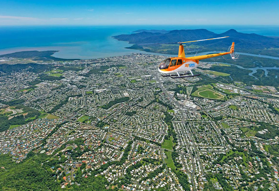 A yellow helicopter flies over a coastal city with dense green areas, residential neighborhoods, and distant mountains near the shoreline under a clear sky.