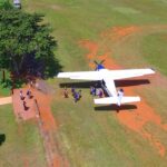 A small propeller plane is parked on a grassy field as a group of people walk from the aircraft toward a fenced area under a tree.