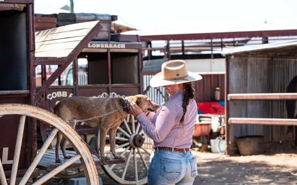 A woman in a hat stands beside a chained dog on a wooden wagon, gently holding the dog's face in an outdoor rural setting.