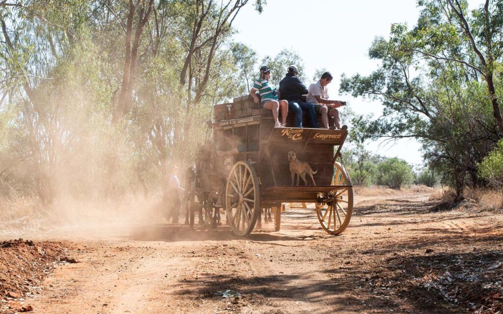 A horse-drawn wooden carriage with passengers rides along a dusty dirt road surrounded by trees. Two dogs sit at the front of the carriage.