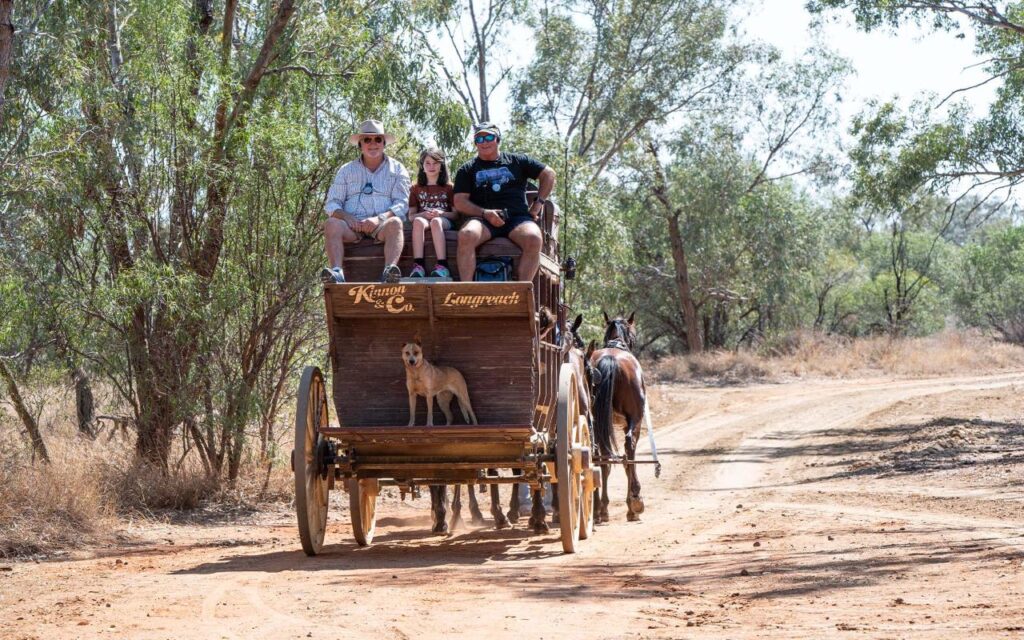 Three people and a dog ride in a horse-drawn wooden wagon on a dirt path surrounded by trees and dry grass.