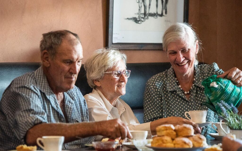 Three older adults sit at a table with tea and scones, one woman pours tea while the others smile and enjoy the conversation.