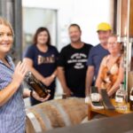 A woman holds a bottle and smiles at the camera in a distillery, with four people standing and smiling in the background near barrels and equipment.