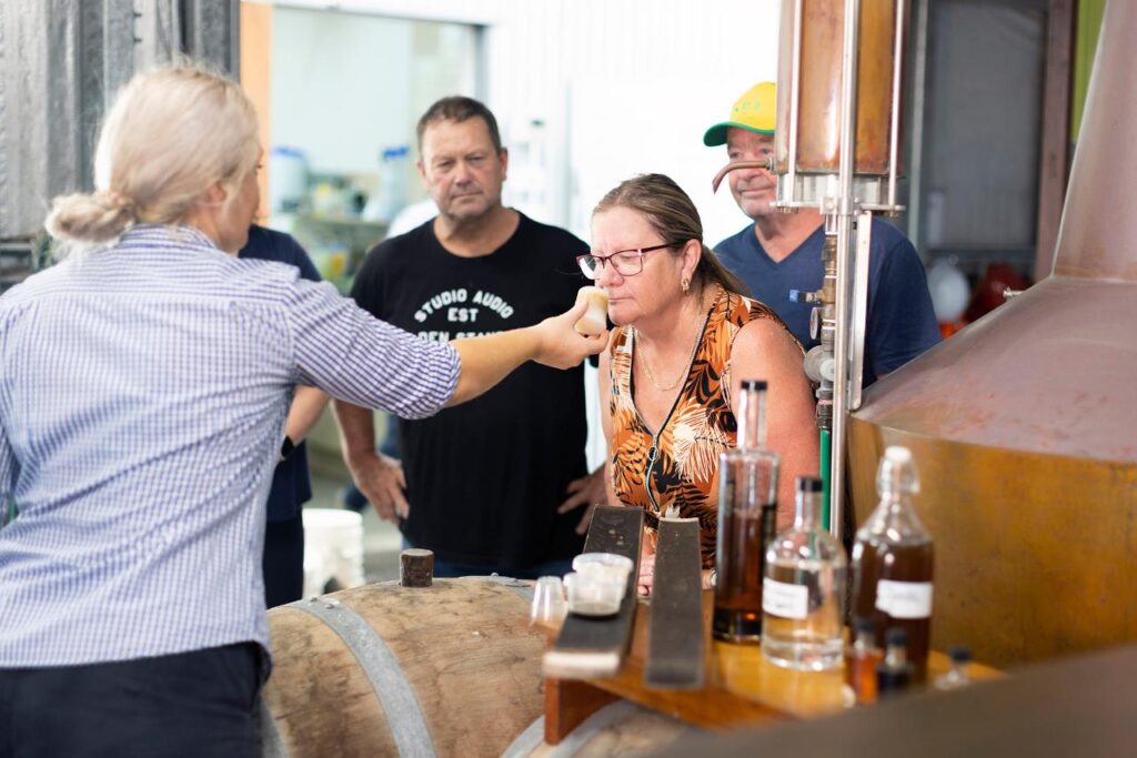 A woman smells the contents of a glass offered by a guide during a distillery tour, while others observe beside a barrel and copper still.