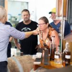 A woman smells the contents of a glass offered by a guide during a distillery tour, while others observe beside a barrel and copper still.
