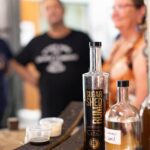 Bottles of rum and spirits on a counter with two tasting cups; two people stand blurred in the background.