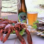 Plates of cooked crayfish and grilled meat skewers are displayed next to a bottle and glass of beer on an outdoor table by the water.