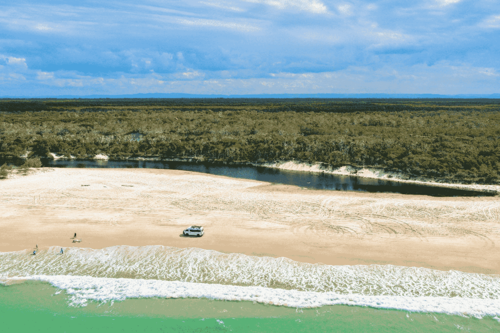 A car is parked on a sandy beach near the shoreline, with a river and forest in the background under a partly cloudy sky.
