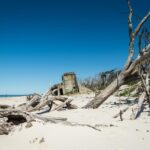 A sandy beach with scattered driftwood and an old concrete structure partially buried in the sand under a clear blue sky.