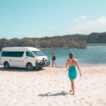 A woman in a turquoise dress walks on sand toward a person standing by a white van near a lake bordered by trees.