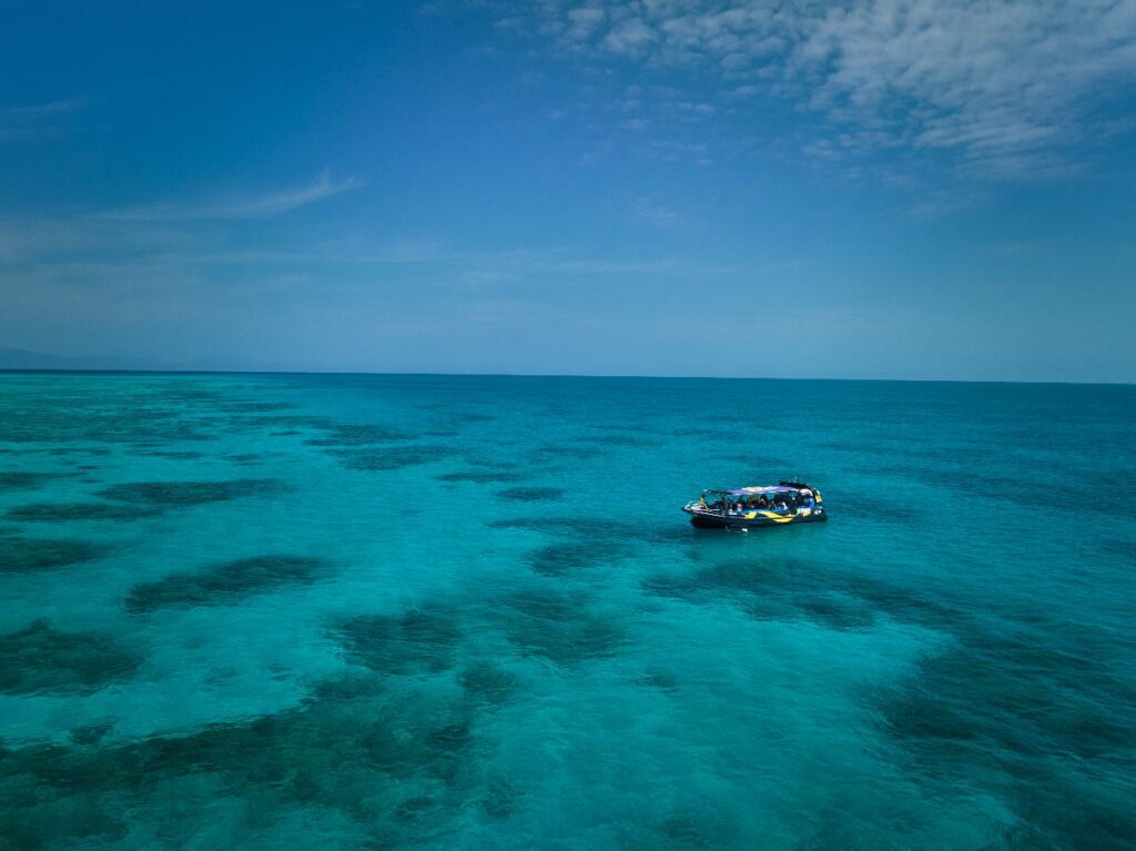 A small boat floats on clear turquoise ocean water with patches of coral visible beneath a partly cloudy blue sky.