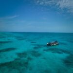 A small boat floats on clear turquoise ocean water with patches of coral visible beneath a partly cloudy blue sky.
