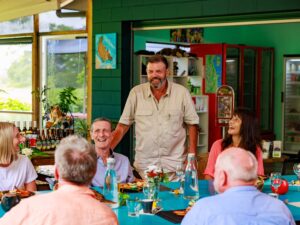 A man stands and speaks at a table, surrounded by five seated adults who listen and smile in a brightly colored room with large windows.