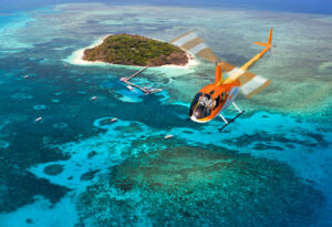 An orange helicopter flies over turquoise waters and coral reefs near a small, green island with a dock and several boats.