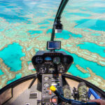 Aerial view from inside a helicopter cockpit flying over turquoise waters and coral reefs, with the pilot’s hands visible on the controls.