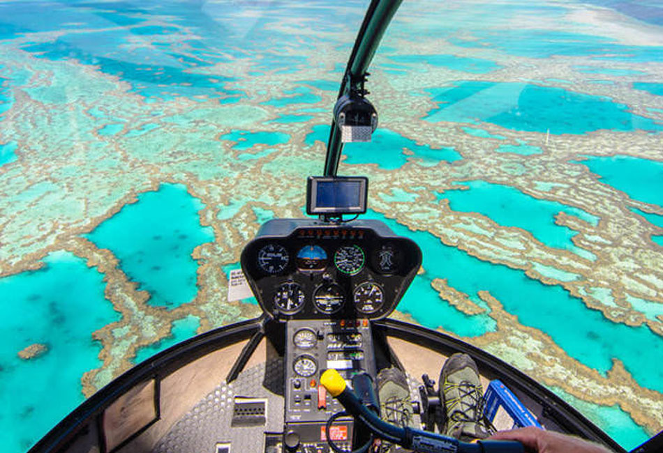 Aerial view from inside a helicopter cockpit flying over turquoise waters and coral reefs, with the pilot’s hands visible on the controls.