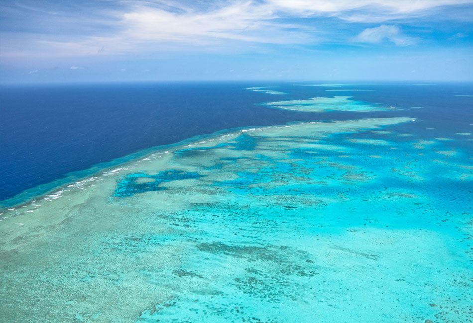 Aerial view of clear turquoise water and coral formations in a large reef system under a blue sky with sparse clouds.