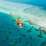 An orange helicopter flies over a coral reef and clear turquoise water near a sandy shoreline.