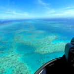 Aerial view from a helicopter cockpit showing clear blue ocean water and coral reefs below under a sunny sky.