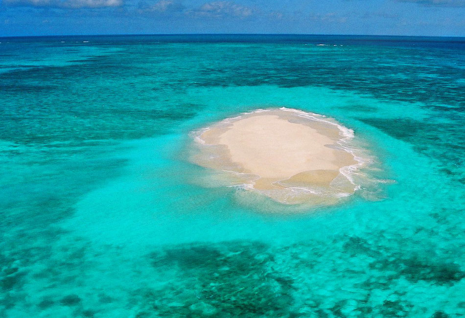 A small, sandy island sits in the middle of clear turquoise ocean water under a blue sky.