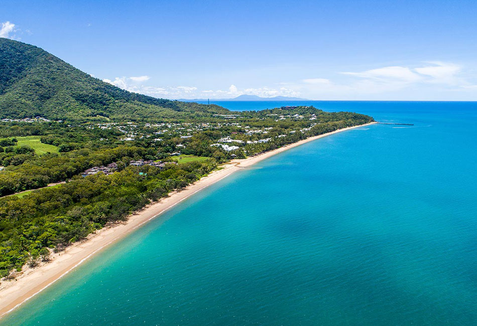 Aerial view of a coastline with turquoise water, sandy beach, green forest, buildings, and a mountain under a clear blue sky.