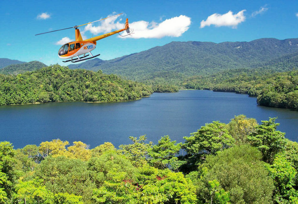 An orange helicopter flies over a lush green forest and a large lake, with mountains and a blue sky in the background.