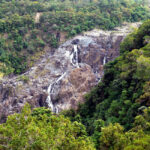 A rocky waterfall with thin streams of water flows down a steep, forested hillside surrounded by dense green trees.