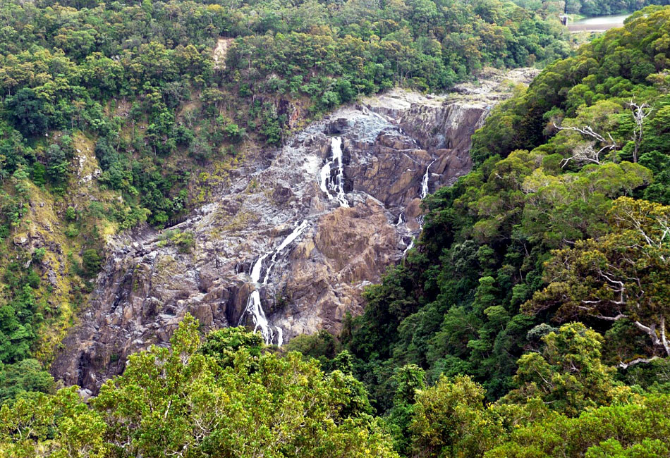 A rocky waterfall with thin streams of water flows down a steep, forested hillside surrounded by dense green trees.