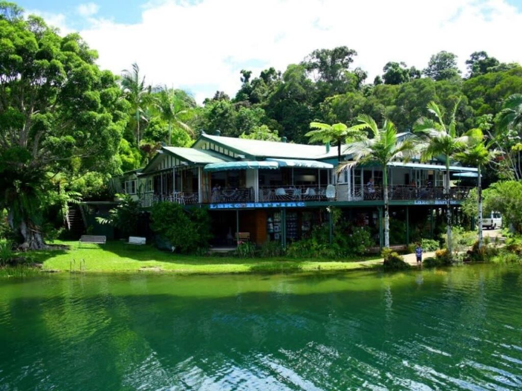 Lakeside building with green roofs and outdoor seating, surrounded by lush trees and reflected in calm water under a partly cloudy sky.