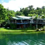 Lakeside building with green roofs and outdoor seating, surrounded by lush trees and reflected in calm water under a partly cloudy sky.