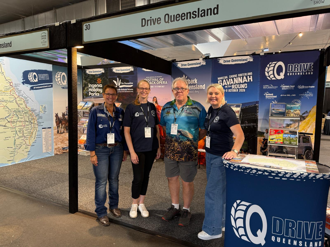 Four people stand smiling in front of a Drive Queensland exhibition booth with maps and promotional banners displayed in the background.