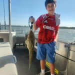 Boy in a red football jersey stands on a boat, smiling and holding up a large fish by the mouth with a grip tool. Water and seats are visible in the background.