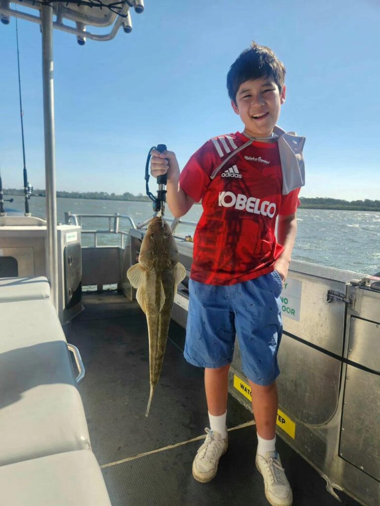 Boy in a red football jersey stands on a boat, smiling and holding up a large fish by the mouth with a grip tool. Water and seats are visible in the background.