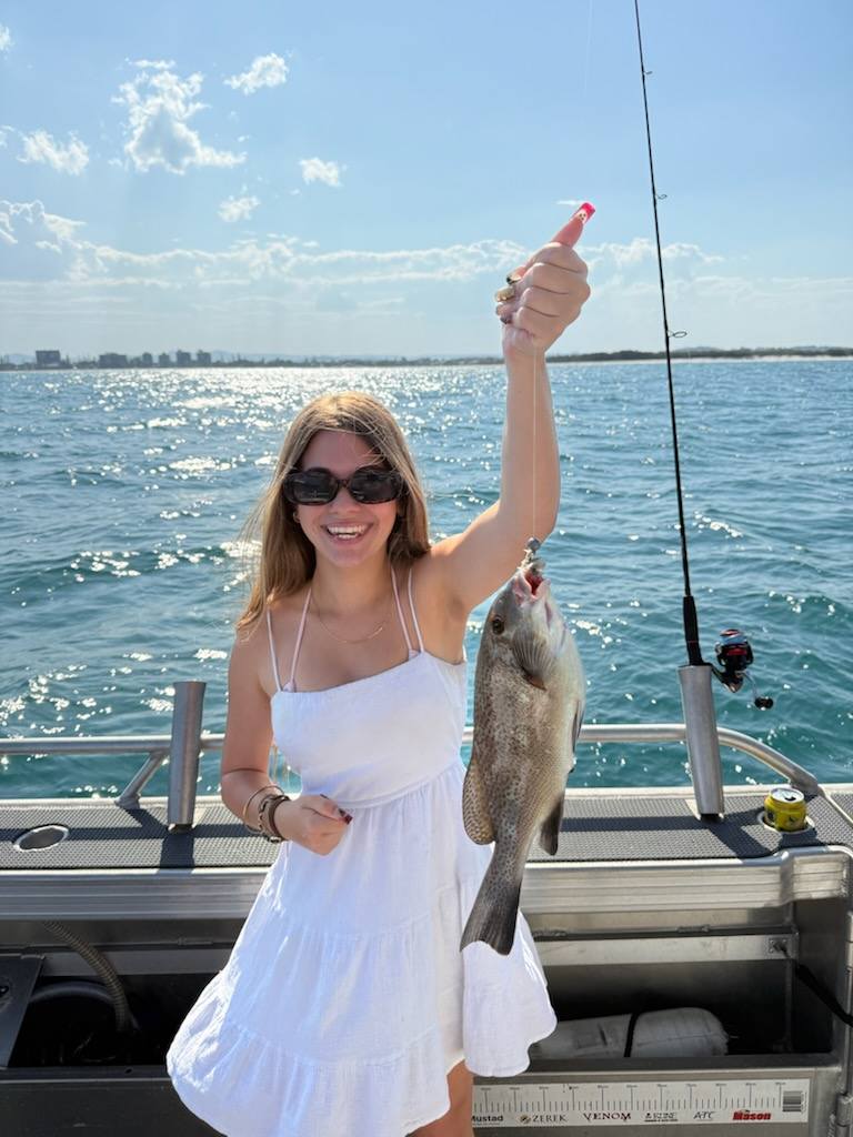 A woman in a white dress stands on a boat holding up a freshly caught fish with one hand; the ocean and distant shoreline are visible in the background.