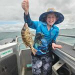 A smiling child on a boat holds up a freshly caught fish with one hand, wearing a blue fishing shirt, shorts, and a wide-brimmed hat.