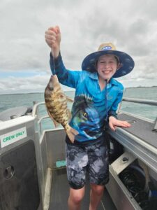 A smiling child on a boat holds up a freshly caught fish with one hand, wearing a blue fishing shirt, shorts, and a wide-brimmed hat.