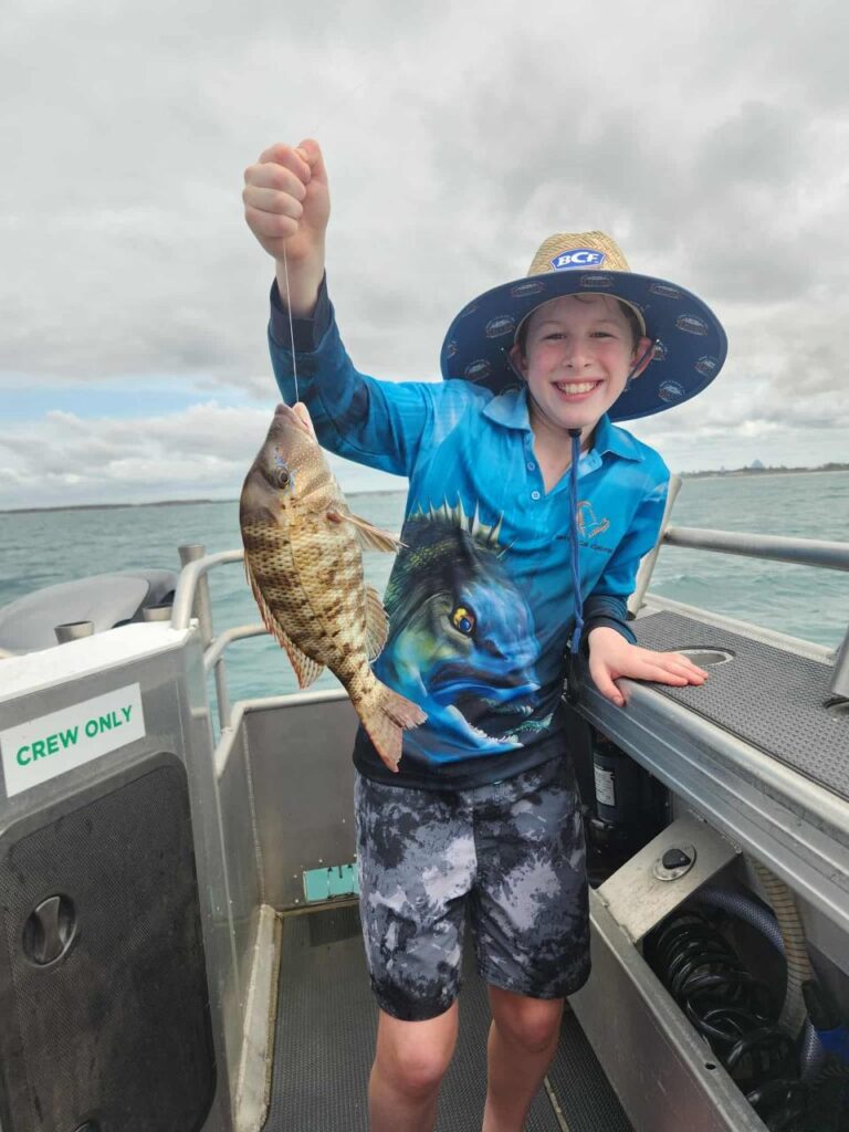 A smiling child on a boat holds up a freshly caught fish with one hand, wearing a blue fishing shirt, shorts, and a wide-brimmed hat.