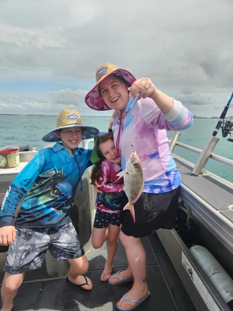 Three people on a boat, one adult holding a small fish on a fishing line, two children stand beside her, all smiling, wearing hats and colorful clothing. Ocean and cloudy sky in background.